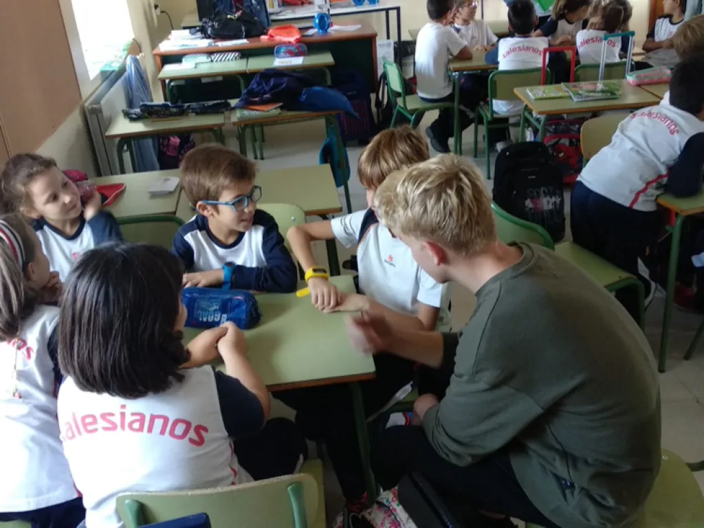 Jugendliche sitzen mit spanischen Schulkindern an Gruppentischen.